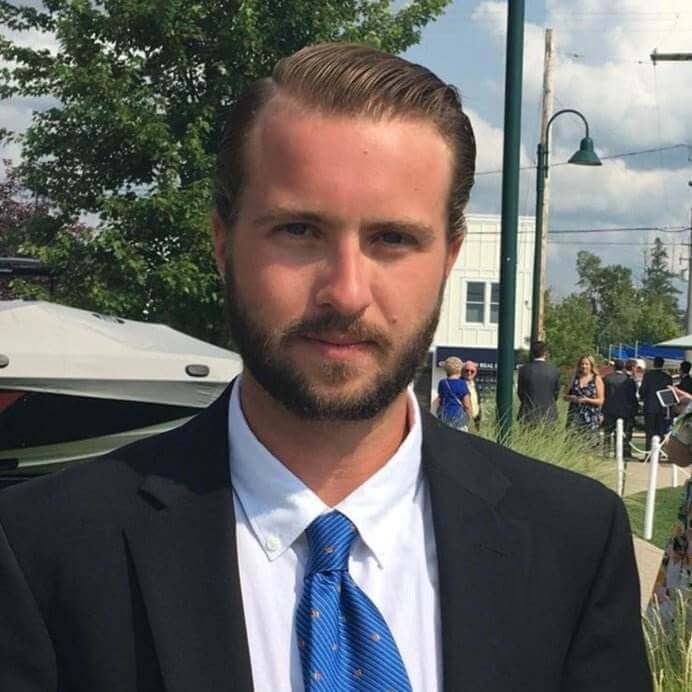 A man with light brown hair and a beard, part of our team, is wearing a black suit jacket, a white dress shirt, and a blue tie. He is standing outdoors under a partly cloudy sky, with trees, buildings, and people in the background.