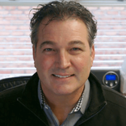 A person with short, dark hair and wearing a dark jacket over a checked shirt is smiling at the camera. In the background, there is a window with blinds and some office equipment, highlighting an environment that understands about safety in the workplace.