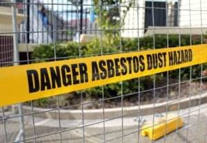 A yellow warning tape with the text "DANGER ASBESTOS DUST HAZARD" is attached to a metal fence, sectioning off an area with blurred foliage and a building in the background. The scene, ideal for blog content, indicates a construction or demolition zone with an asbestos risk.