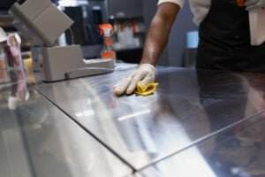 A person wearing a white shirt, black apron, and plastic gloves cleans a stainless steel surface with a yellow cloth. In the background, cleaning supplies and a dark-colored counter can be seen, ready for the next task. Perfect for your blog on efficient cleaning tips.