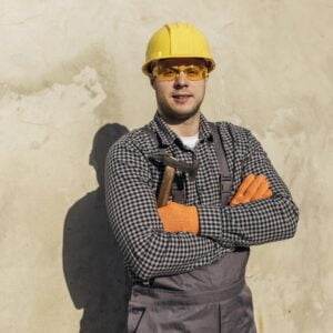 A man wearing a yellow hard hat, yellow safety glasses, orange gloves, a checkered shirt, and gray overalls is standing against a beige wall. With his arms crossed and holding a hammer, he exemplifies construction safety while looking confident and prepared.
