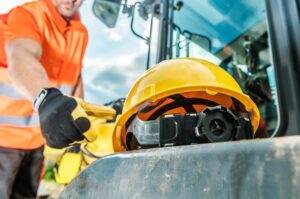A construction worker in an orange safety vest reaches toward a yellow hard hat resting on a piece of machinery. The scene includes a reflective glove and a partially visible vehicle, emphasizing the importance of construction safety in this industrial setting.