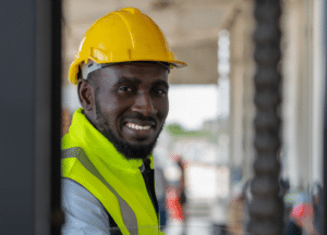 A man wearing a yellow hard hat and a high-visibility safety vest smiles at the camera. He is standing in an industrial setting with blurred background elements, possibly indicating a construction site or factory where federal OSHA regulations are likely enforced.