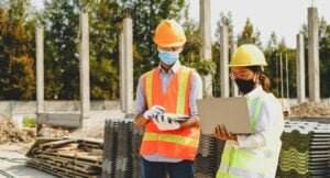 Two construction workers wearing orange safety vests, hard hats, and face masks are on a construction site. One worker is looking at a tablet while the other holds a laptop. In the background, there are building materials and half-built structures—a scene exemplifying Safety Meeting Basics in action.