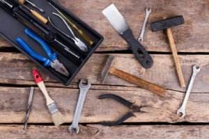 A variety of tools are arranged on a rustic wooden surface, including pliers, wrenches, screwdrivers, a paintbrush, hammers, and other hand tools. Hand tool safety tips are essential when using these items. A black toolbox partially visible in the top left corner holds more tools.