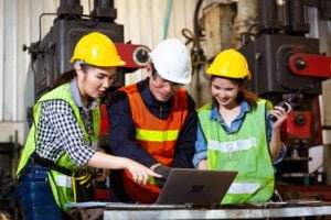 Three construction workers in safety gear and hard hats gather around a laptop in a factory setting, likely reviewing a safety program. One points at the screen, another holds a walkie-talkie, and the third observes. Machinery is visible in the background.