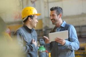 Two men in a workshop smile and talk. One wears a yellow hard hat and gray work jacket, while the other, in a blue shirt and gray vest, holds a tablet, gesturing towards the screen. The background shows industrial equipment and a blurred workshop setting, emphasizing their focus on the safety program.
