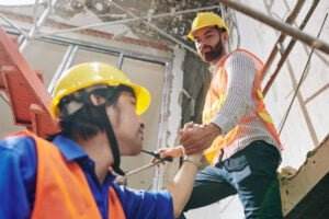 Two construction workers wearing hard hats and safety vests, part of a comprehensive safety program, are on a building site. One worker, positioned on a higher platform, is helping another climb up by holding his hand. The background shows scaffolding and a partially constructed structure.