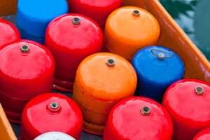 A collection of brightly colored gas cylinders, primarily red with a few orange and blue ones, neatly arranged in a wooden container. The reflective surfaces and valve tops emphasize the precautions necessary for handling these potential combustible liquids.