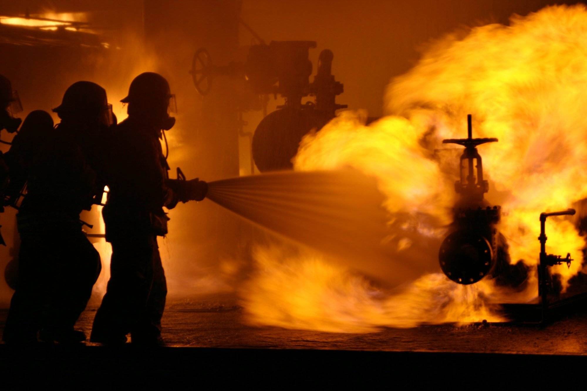 Firefighters in protective gear battle a large industrial fire, with one spraying water onto the flames. Practicing rigorous fire safety measures, they work tirelessly as the intense orange blaze engulfs machinery in the background, casting a bright glow and creating thick smoke.