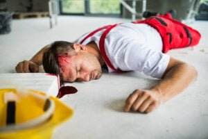 A man in red overalls and a white shirt is lying unconscious on the floor with a head injury, bleeding from his forehead. A yellow caution sign is visible nearby, indicating the plant accidents that often occur in this hazardous environment.