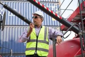A man wearing a reflective safety vest, hard hat, and safety sunglasses communicates using a walkie-talkie. He is standing by industrial machinery and shipping containers in the background. The setting appears to be a construction or shipping site.