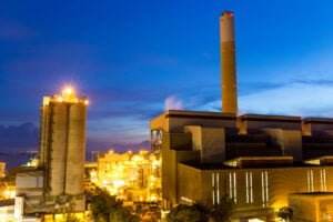 A brightly lit industrial power plant, specializing in concrete manufacturing, features various structures and a tall smokestack emitting a small amount of smoke, set against a deep blue twilight sky. The illuminated buildings and equipment create a striking contrast against the evening backdrop.