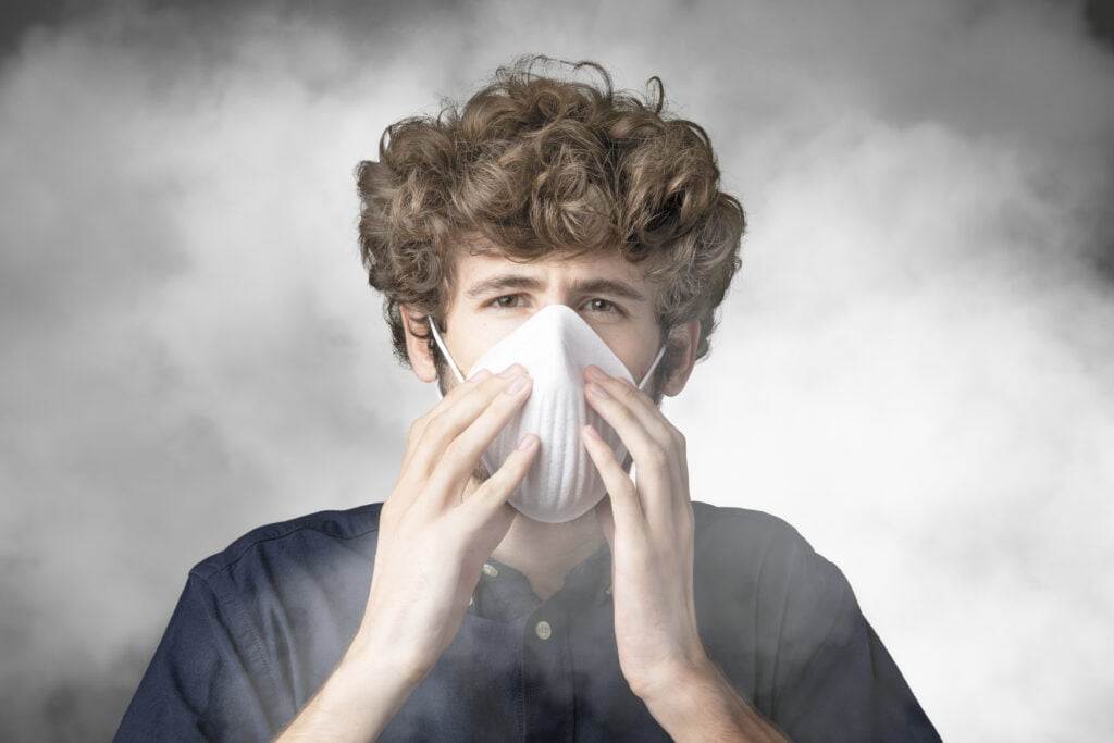 A young person with curly hair wearing a blue shirt and head protection is covering their mouth and nose with a white mask. The background is filled with smoke or fog, creating a hazy effect. The person looks directly into the camera, appearing concerned.