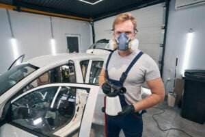A man in a grey t-shirt and blue overalls, wearing black gloves and respiratory protection, stands in a garage holding a paint sprayer. A white car with its door open is visible behind him.