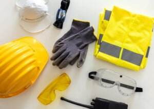A flat lay of protective equipment, including a yellow hard hat, a pair of grey gloves, a yellow high-visibility vest, a dust mask, yellow safety goggles, clear protective goggles, a headlamp, and a walkie-talkie.