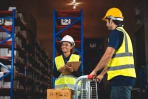 Two workers in safety vests and helmets are seen in a warehouse. One person is holding a clipboard and smiling, while the other is pushing a cart with boxes. Shelves filled with supplies are visible in the background, reflecting the effectiveness of their safety program.