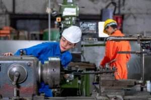 Two workers in a machine shop, both wearing safety helmets and protective gear. In the foreground, a person in a blue uniform operates a lathe machine, with guarding considerations clearly implemented. In the background, a person in an orange uniform works on another machine. The setting is industrial.