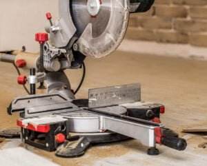 A close-up of a miter saw placed on a workbench. The saw has measurements marked on its base and red adjustment knobs. The blade guard is partially open, revealing a circular saw blade. Machine guards and sawdust are scattered around the tool, indicating recent use.