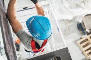 An overhead view of a construction worker wearing a blue hard hat and a safety harness, diligently working on scaffolding at a busy construction site. Various equipment and materials are scattered on the ground below, while birds and other flying objects move through the scene.
