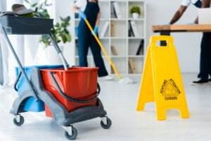 A mop bucket on wheels with red and blue sections is in the foreground. A yellow caution sign is nearby. In the background, two people are cleaning an office: one is sweeping the floor, while the other wipes a table. Shelves with various items are visible in the room.