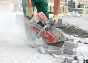 A worker wearing green pants and gloves operates a large red concrete saw to cut gray bricks or pavers on a construction site. Dust surrounds the area as the saw cuts through the material, raising concerns about silica exposure, and stacks of bricks are visible in the background.