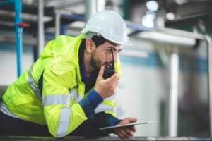 A man wearing high-visibility clothing, including a yellow jacket and a white hard hat, is crouching and speaking into a walkie-talkie. He holds a clipboard in his other hand and appears to be in an industrial environment.