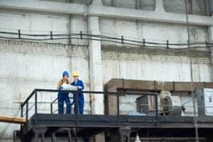 Two industrial workers in blue coveralls and safety helmets stand on a black metal platform equipped with fall protection in a factory. They are looking at a large sheet of paper, possibly a blueprint. The background reveals a large, high-ceilinged industrial space with various machinery.
