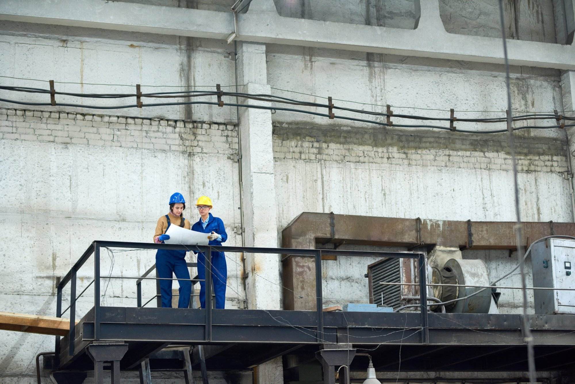 Two industrial workers in blue coveralls and safety helmets stand on a black metal platform equipped with fall protection in a factory. They are looking at a large sheet of paper, possibly a blueprint. The background reveals a large, high-ceilinged industrial space with various machinery.