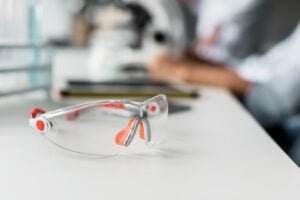 A pair of transparent safety glasses with orange ear guards rests on a white tabletop. In the background, a blurred figure operates a microscope, suggesting a laboratory environment where safety meetings are likely conducted.