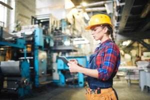 A worker in a plaid shirt and yellow hard hat stands in an industrial setting, using a tablet. Machinery and equipment are visible in the background. The worker is also wearing safety glasses and has a tool belt around their waist, taking every precaution to avoid an accident.