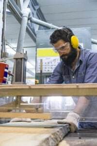 A man with a beard wearing safety goggles, ear protection, and gloves is operating a table saw fitted with machine guards in a workshop. He is focused on cutting a piece of wood, and industrial equipment is visible in the background.