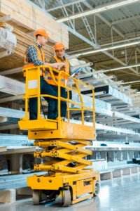 Two construction workers wearing orange helmets and safety vests are standing on a yellow scissor lift, examining documents. They are in a large warehouse with metal beams and building materials on shelves in the background, meticulously planning their next task.