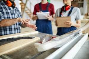 Three people in a workshop, two wearing denim overalls and red shirts, listening to a person gesturing about table saw safety. They have safety earmuffs and hold notepads. Various woodworking tools and a circular saw are on the table in front of them.