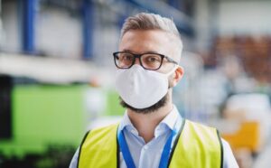 A man with grey hair and a beard, wearing glasses and a white face mask, is pictured in an indoor industrial setting. He is dressed in a yellow safety vest over a light blue shirt, holding an ID card on a blue lanyard around his neck, possibly working with acetone.