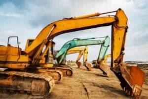 A lineup of colorful excavators, including yellow, blue, and green, stand idle on a sandy construction site under a partly cloudy sky. Used for trenching and excavating, the machines have large, rusted shovels and are arranged in a row, with tracks imprinted in the sand beneath them.