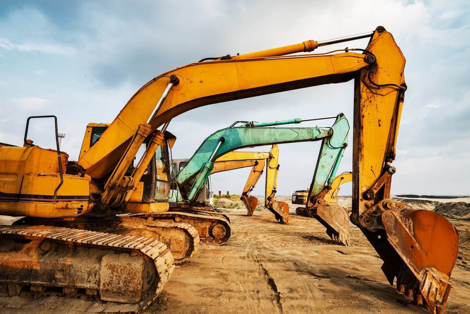 A lineup of colorful excavators, including yellow, blue, and green, stand idle on a sandy construction site under a partly cloudy sky. Used for trenching and excavating, the machines have large, rusted shovels and are arranged in a row, with tracks imprinted in the sand beneath them.