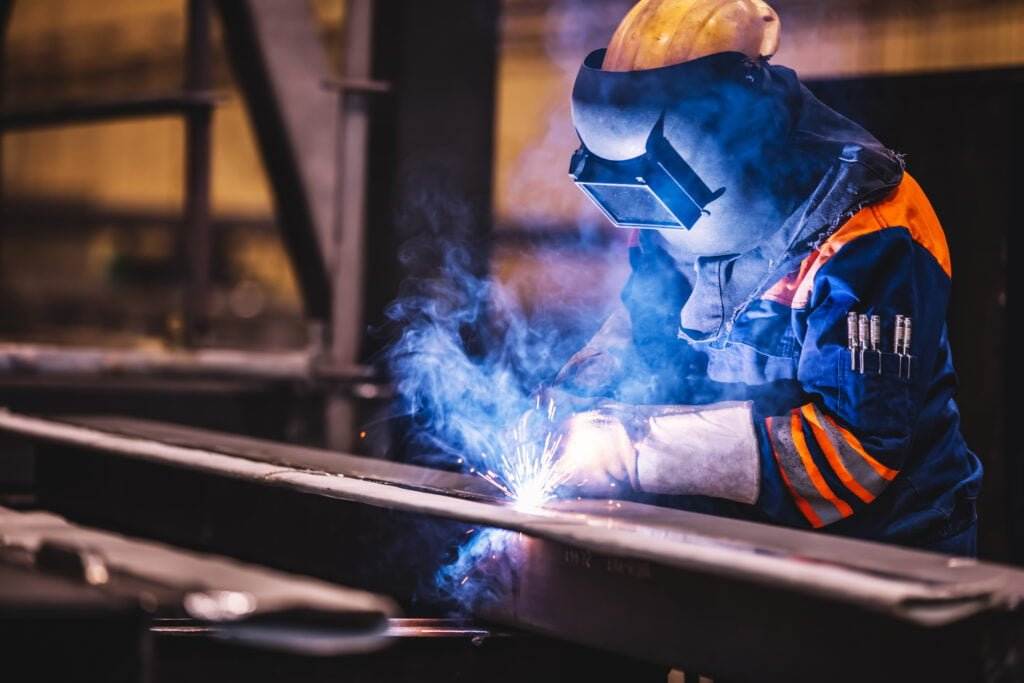 A worker in protective gear, including a welding helmet and gloves, welds metal in a workshop. Sparks and blue smoke surround the welding site, emphasizing the importance of welding and cutting safety. The environment appears industrial with metal structures and equipment in the background.