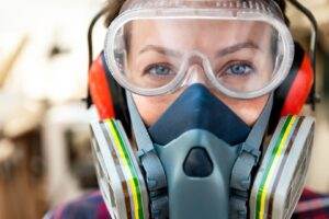 Close-up of a person working safely with lead, wearing protective gear including a respirator mask, safety goggles, and earmuffs. Their eyes are visible through the goggles against a blurred background, suggesting they are in a work environment.