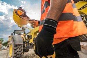 A construction worker, donned in a fluorescent orange safety vest and black gloves, holds a hard hat near a yellow construction vehicle. The worker's smartwatch is visible on their wrist, highlighting the importance of monitoring for heat illness. The scene is outdoors with a cloudy sky in the background.