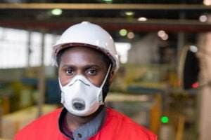 A worker wearing a white hard hat and a protective face mask stands in a factory setting, potentially preventing hearing loss. He is dressed in a red work jacket. The background is blurred, showing an industrial environment with various equipment and machinery.