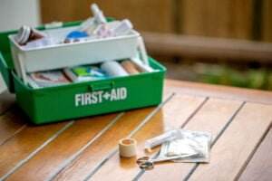 A green first aid kit labeled "FIRST + AID" is open on a wooden surface, revealing various medical supplies inside. In the foreground, among the scattered items including scissors, a syringe, adhesive tape, and other small medical items, there's an ear plug case addressing hearing loss needs.