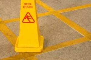 A yellow caution sign with "Caution Wet Floor" written on it and a slipping person icon is placed on a concrete floor with yellow grid lines. Though the surface appears dry, those with hearing loss might miss verbal warnings, making the visually distinctive pattern around the sign crucial for their safety.