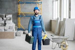 A construction worker wearing a blue hard hat and overalls smiles while standing in a partially constructed building. He holds a toolbox and a bucket, with a wheelbarrow, some insulation panels, and a stack of bricks in the background. Scaffolding is also visible, demonstrating safe housekeeping on-site.