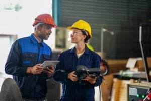 Two industrial workers in protective gear stand in a workshop. The man in the red hard hat holds a tablet and speaks to the woman in the yellow hard hat, who is holding a power tool. Emphasizing the need to avoid safety complacency, they discuss protocols amidst machinery and equipment.