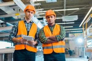 Two workers in a well-lit warehouse are wearing orange safety vests and hard hats, emphasizing safety in the workplace. One holds a tablet while the other stands with arms crossed. Shelves and industrial equipment fill the background, highlighting their organized work environment.
