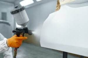 A person wearing orange gloves and white protective gear is using a spray gun for spray painting a white object in a workshop environment. The background is blurred, focusing on the painting process.