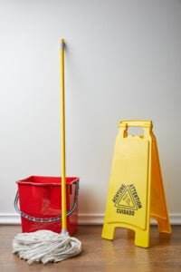 A yellow wet floor sign stands next to a red mop bucket with a yellow mop inside it, leaning against a white wall on a wooden floor. The wet floor sign, designed to prevent falls, features "CUIDADO," "ATTENTION," and "AVERTISSEMENT," indicating caution in multiple languages.