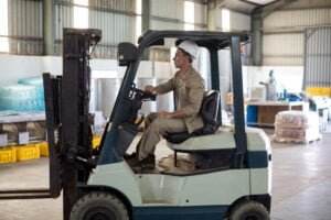 A person wearing a hard hat and beige work clothes operates a forklift inside a spacious warehouse, prioritizing forklift safety. Various items, including crates and equipment, are stacked and organized in the background.