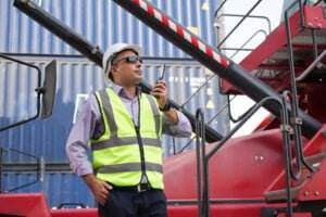 A man wearing a safety vest, hard hat, and sunglasses is using a walkie-talkie. He is standing next to industrial machinery and shipping containers, indicating a construction or industrial site. The scene is outdoors during the day, with his sunglasses shielding him from harsh UV rays.